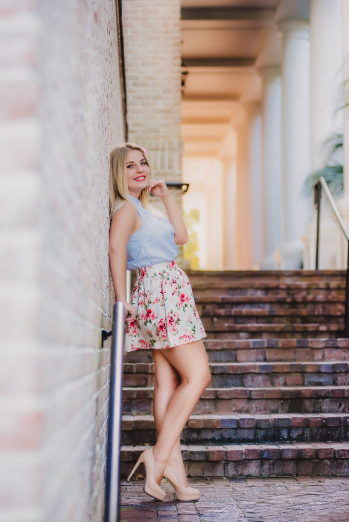A high school senior girl in a floral skirt is leaning on the wall with beautiful golden lighting in the background