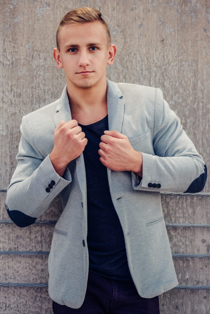 A high school senior boy in a gray blazer leaning against a wall.