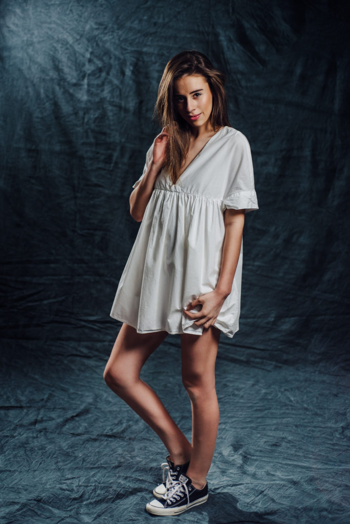 Studio portrait of high school senior girl in a white dress posing on a dark background.