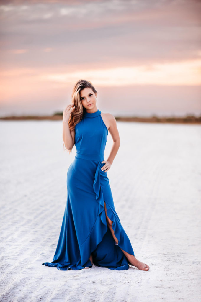 A beautiful high school senior girl in a blue dress posing in the sand at sunset.