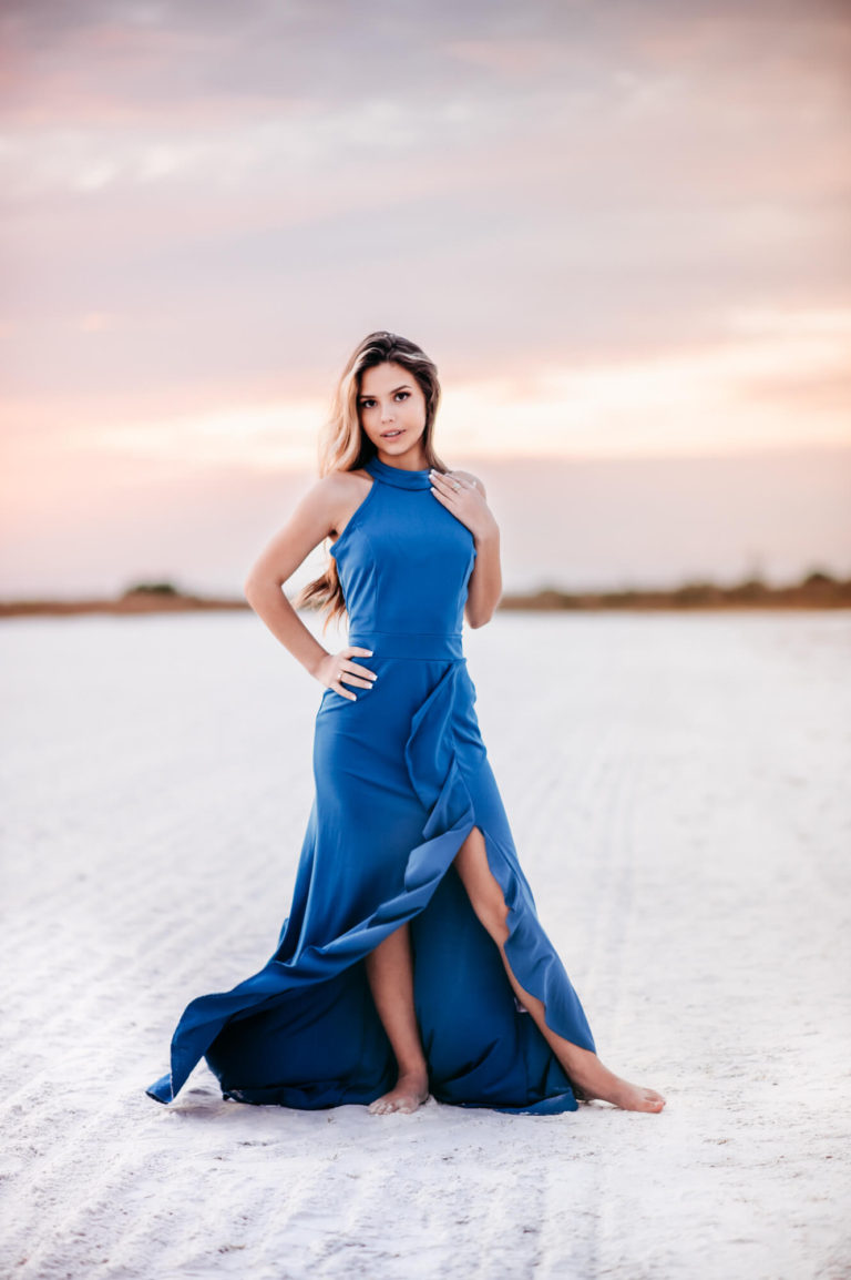 A beautiful high school senior girl in a blue dress standing in the sand on Siesta Key Beach at sunset.