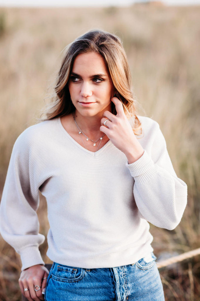 A close up of high school senior in jeans and a white sweater standing in a field.