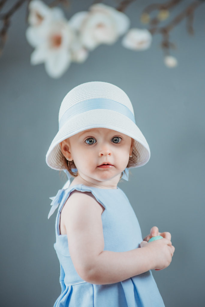 A little girl in a blue dress with a white hat looking at the camera with her beautiful blue eyes