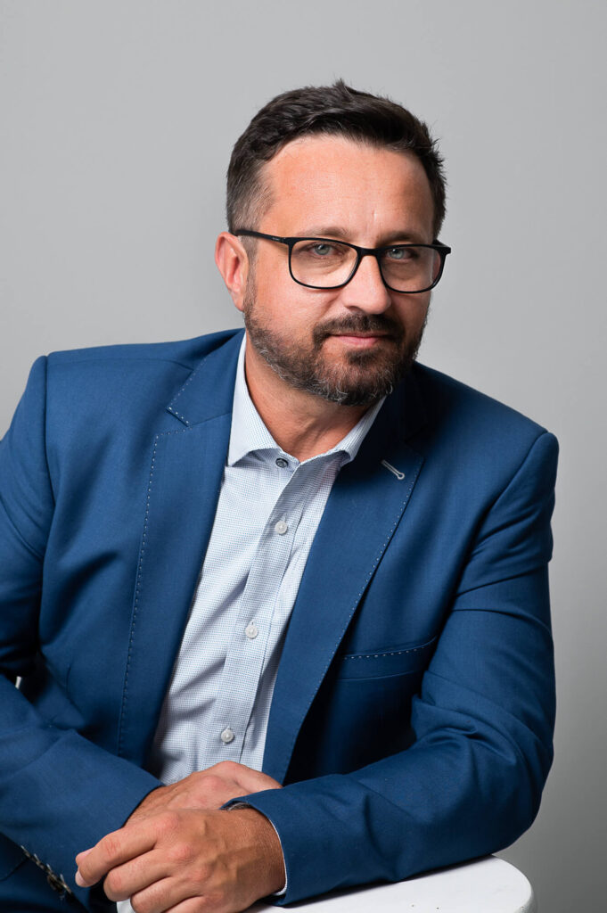 Modern headshot of the men in  light blue shirt and blue jacket wearing  glasses leaning on the table  on light background