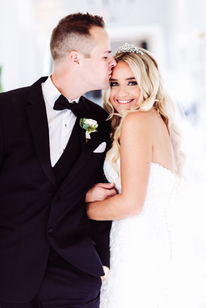 A groom kissing bride forehead when she looks straight into camera smiling in hall of Sarasota Wedding and Golf Venue
