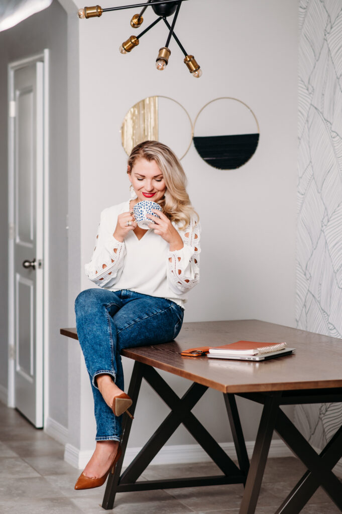 A woman posing for her personal brand photoshoot while sitting on a wooden table with a cup of coffee.