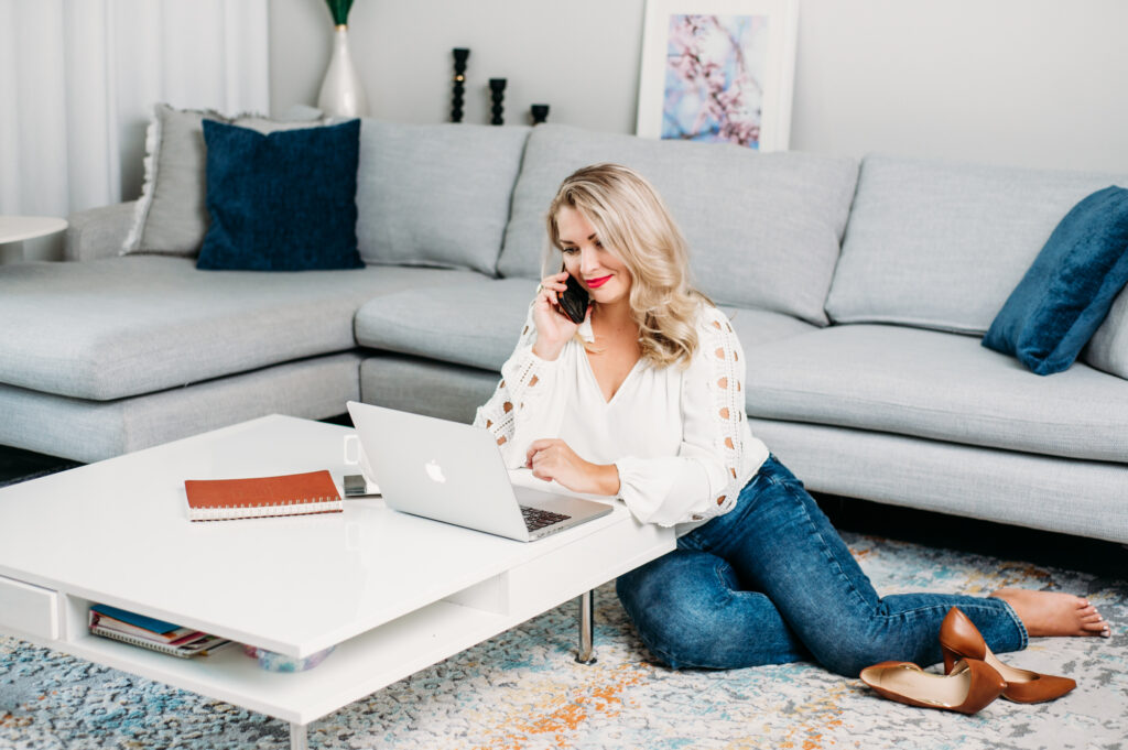 A woman sits on the floor, working on her laptop while speaking on the phone.