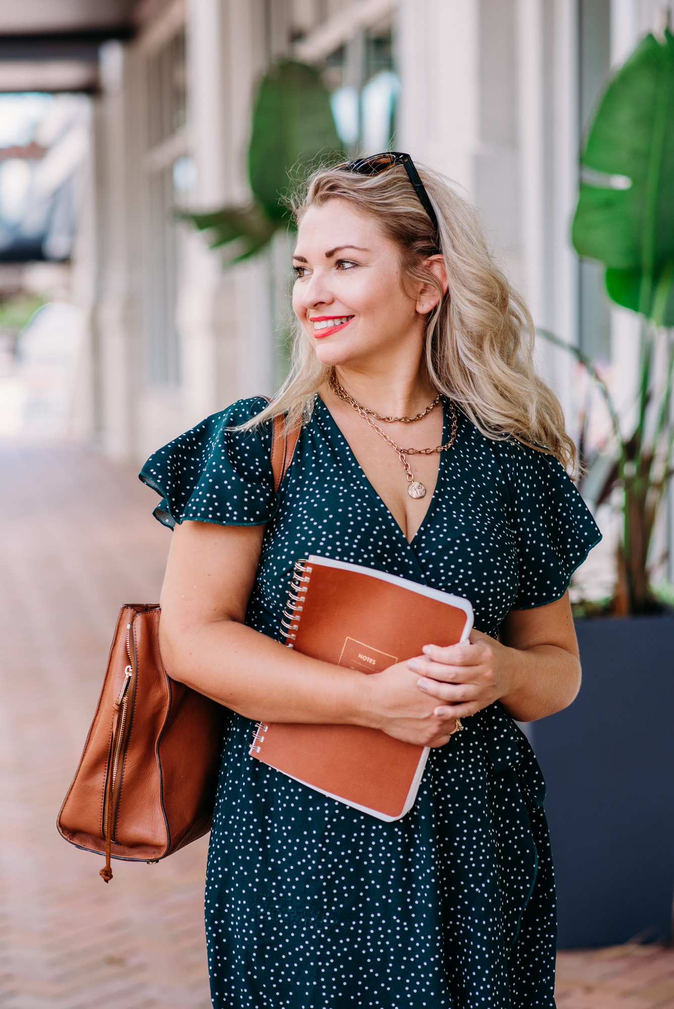 A personal branding image of a life coach strolling down city streets, dressed in a green polka dot dress, holding a brown journal, smiling and glancing to the side.