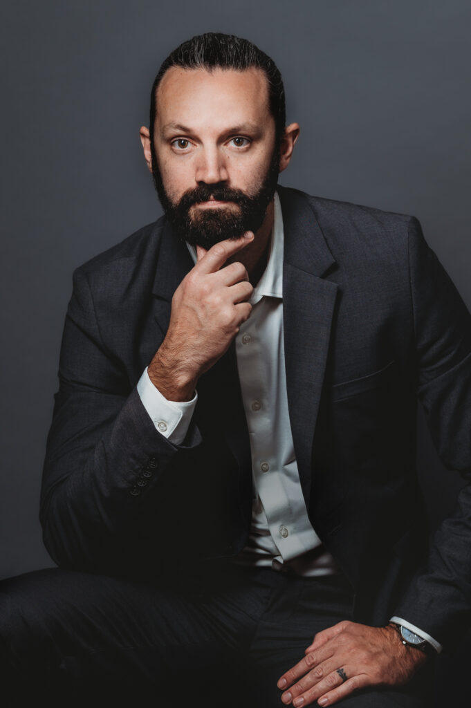 A sophisticated man in his dark suit against a dark grey backdrop. His intense brown eyes lock onto the camera, The well-groomed beard adds an air of rugged refinement to his captivating presence.