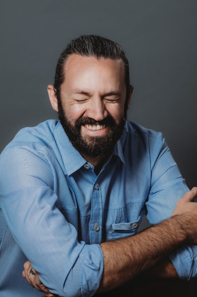 This contemporary portrait captures a man's modern allure as he sports a blue shirt against a backdrop of understated grey. His genuine smile and inviting brown eyes create a welcoming atmosphere.