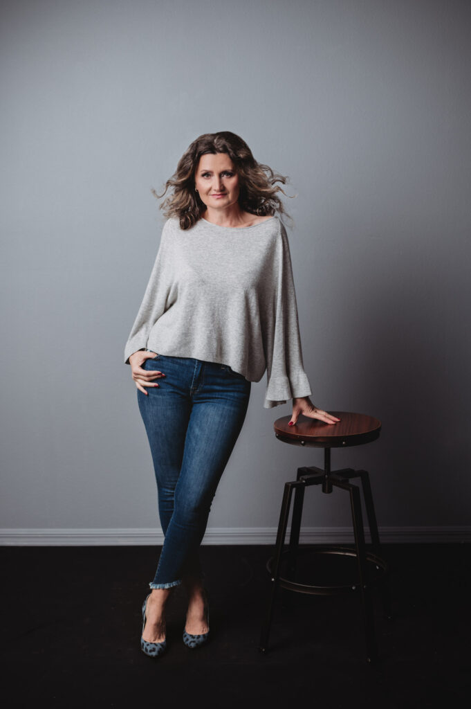 A woman in jeans and a grey sweater striking a pose at a portrait studio for her personal branding photoshoot