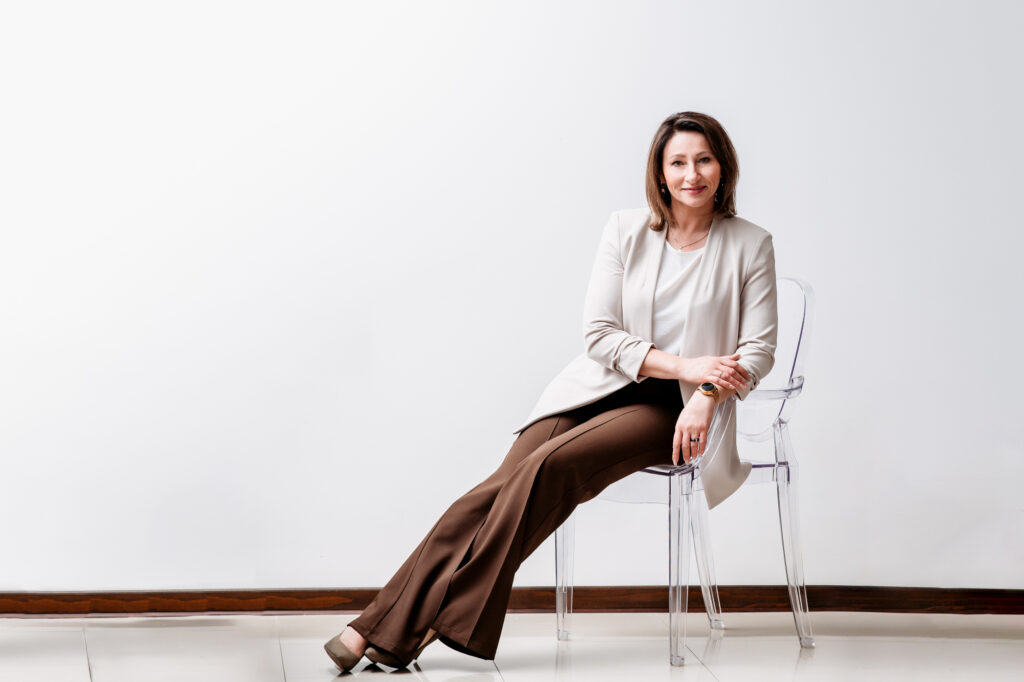 A professional woman in light blazer and brown pentssitting on a clear chair for a business portrait.