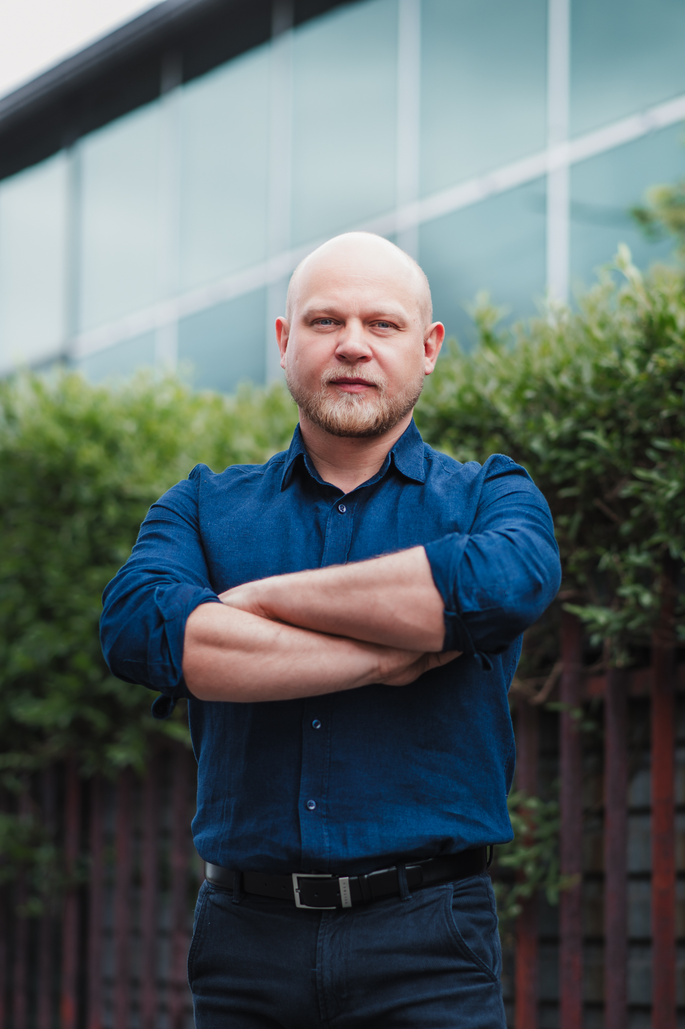 A professional portrait of a bald man in a blue shirt standing in front of a building.