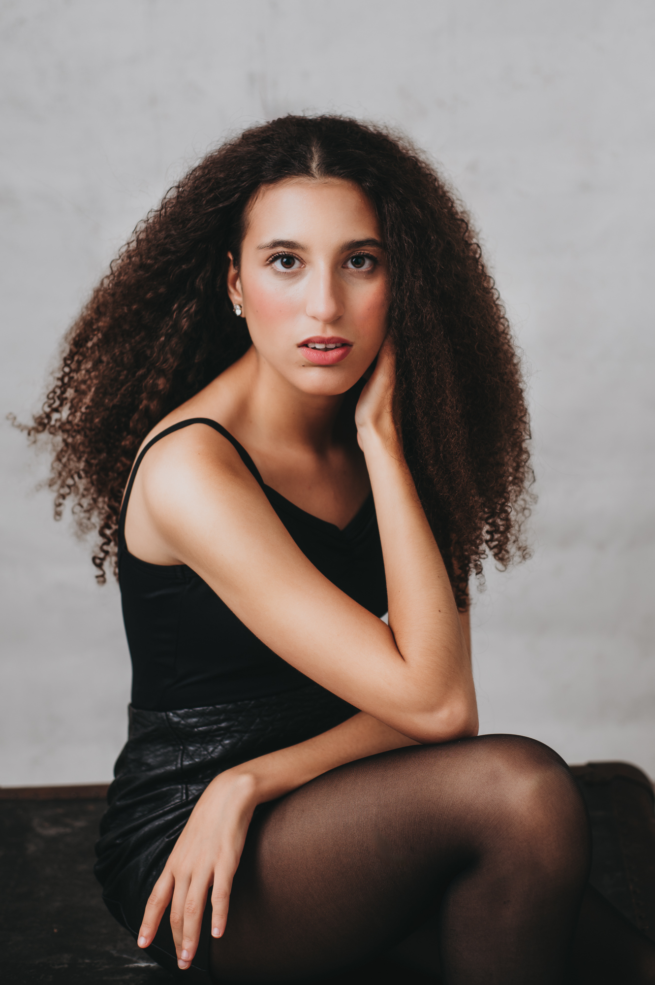 Modern headshot of A young woman with curly hair sitting on a chair.