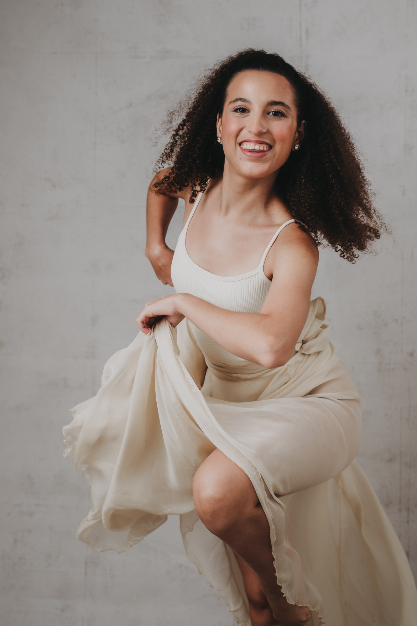 A young woman in a white dress is dancing in front of a gray background.