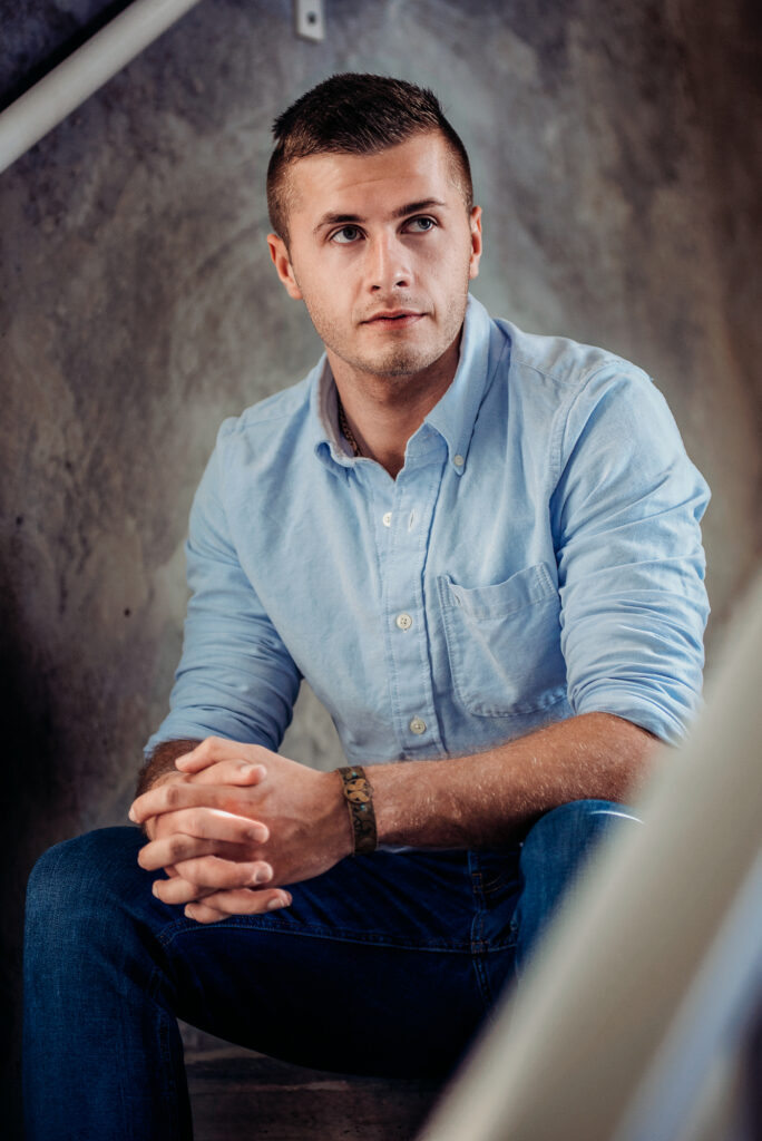 A senior man in a blue shirt sitting on a stair, photographed for his senior portrait.