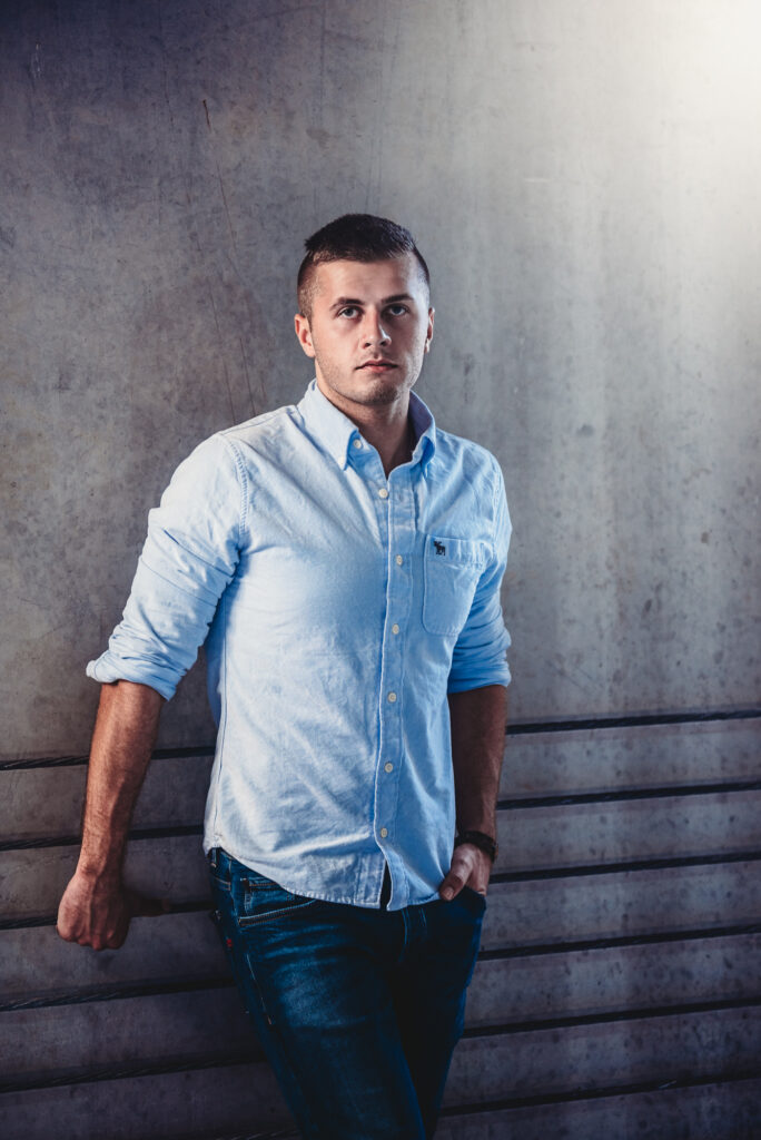 A man in a blue shirt posing for his senior portrait leaning against a wall.