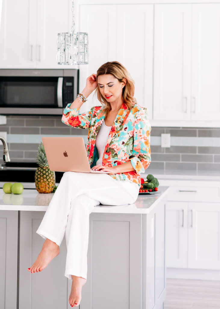 Lifestyle portrait of Health coach siting on the countertop at bright light kitchen working on her laptop wearing colorfull blazer and white pants