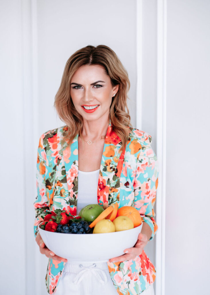 Brand photo of the women in flowery jacket holding a bowl full of broccoli, carrots, oranges, strawberries lemon and apples