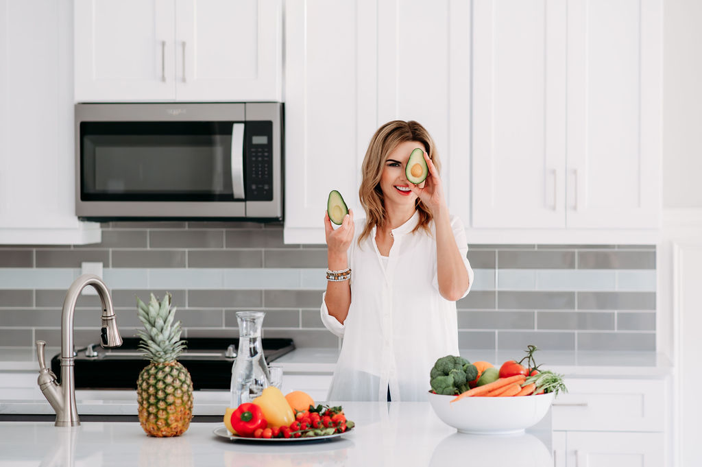 Branding photo of health coach in the kitchen with trays of fruits and veggie holding avocado in her hands