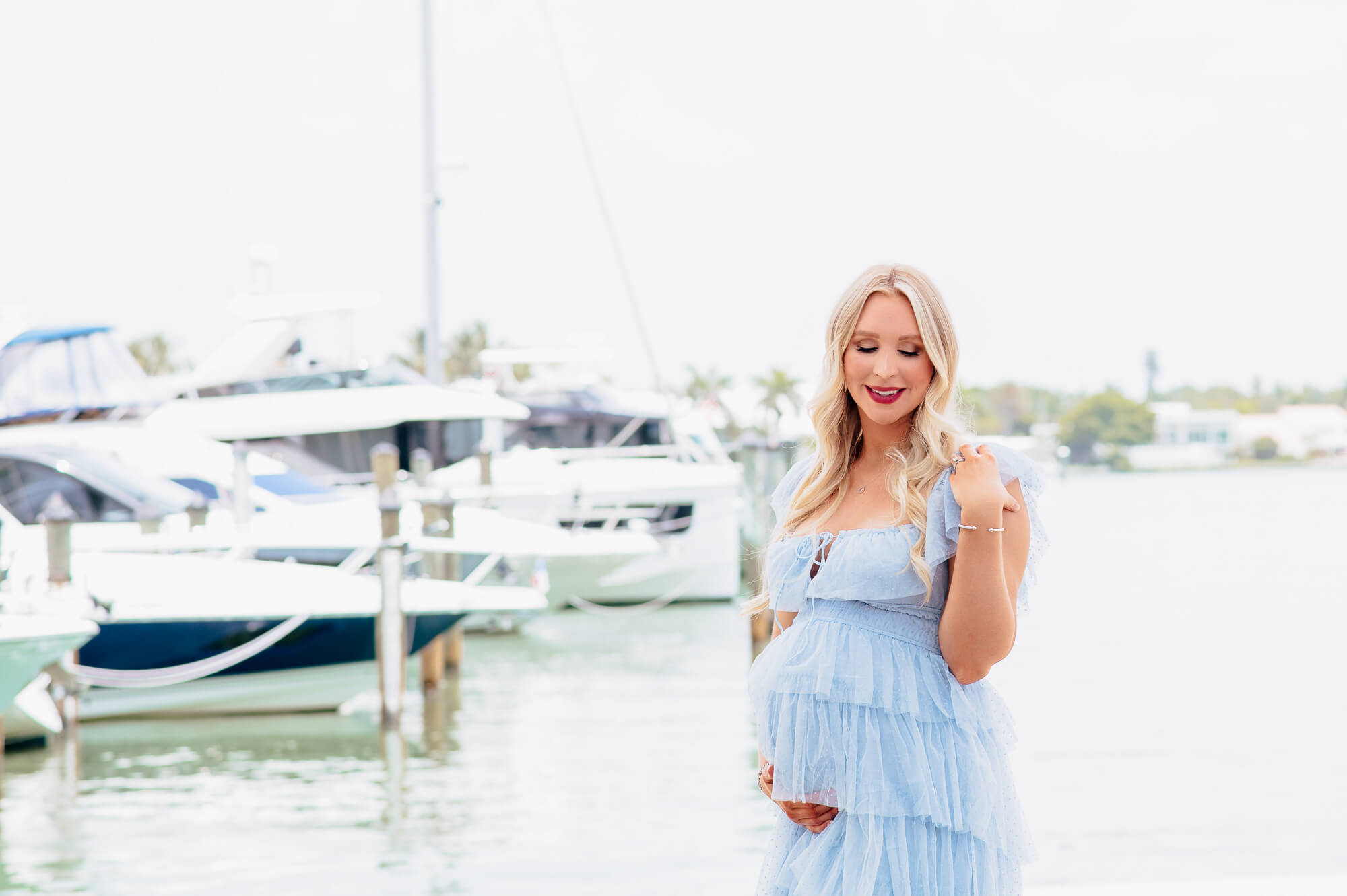 A breathtaking outdoor portrait of a maternity client, wearing a light blue dress, gazing lovingly at her belly with the picturesque Sarasota Yacht Club in the background.