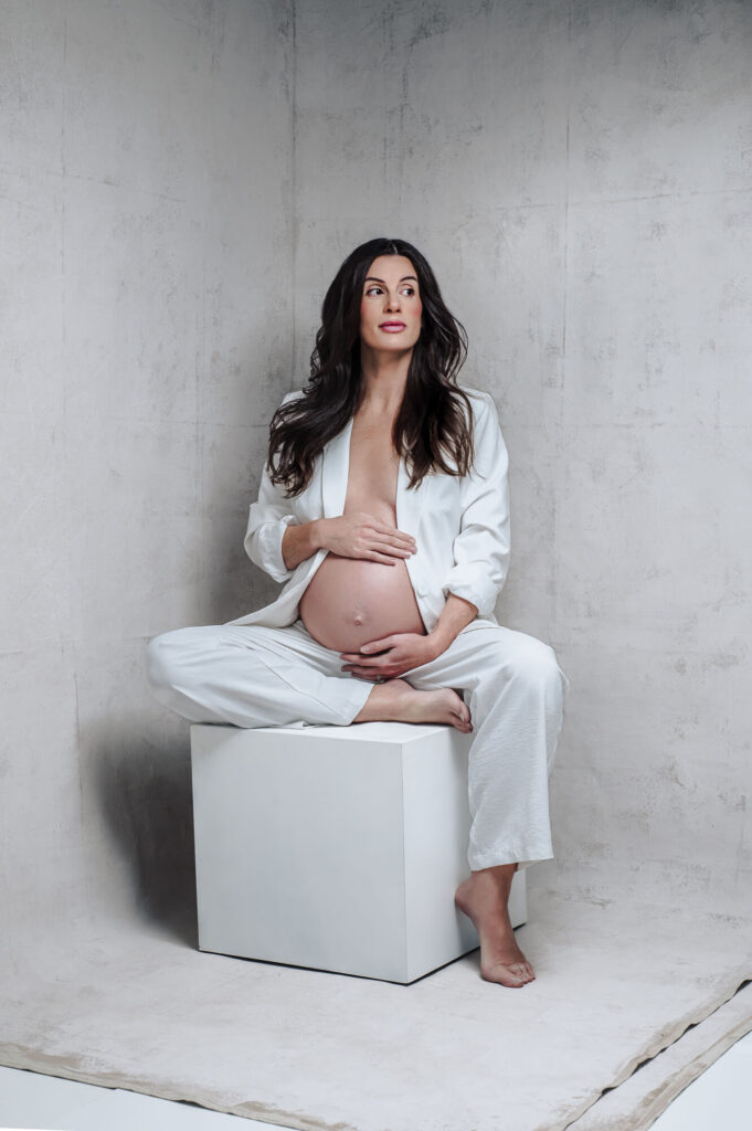 Modern studio portrait of maternity client wearing white suit siting on white block and looking on the side