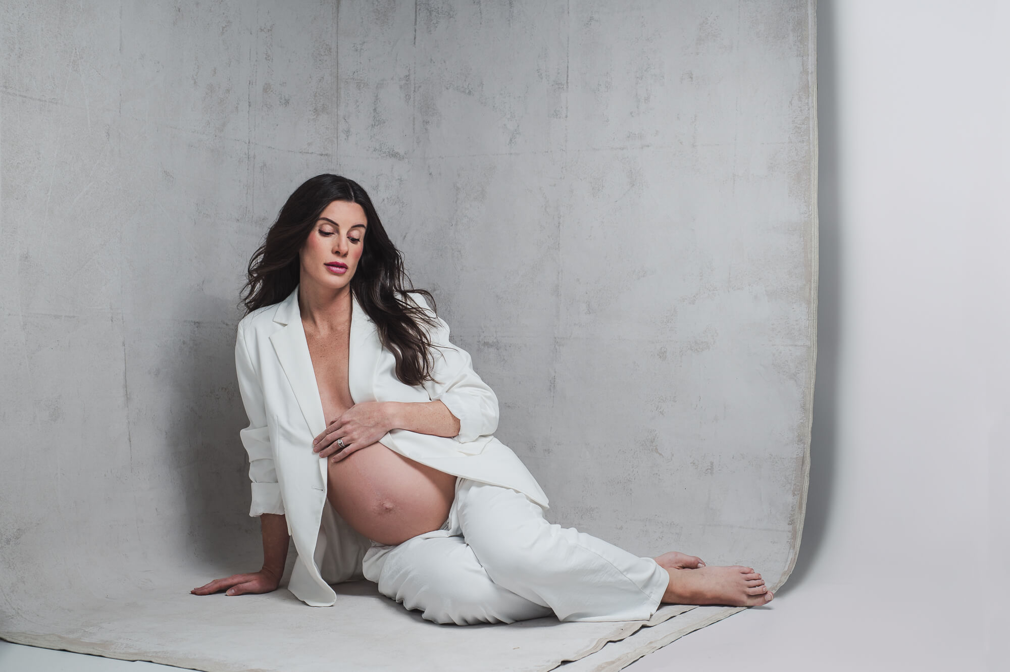 A contemporary studio portrait features a pregnant woman in a white suit, seated on the floor as she lovingly gazes at her belly.Maternity Portrait