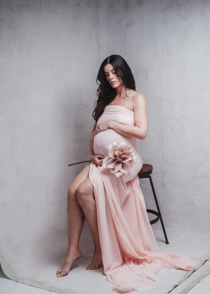 Intimate portrait of dark hair pregnant women with close eyes wrap in peach color fabric holding a flower next to her belly