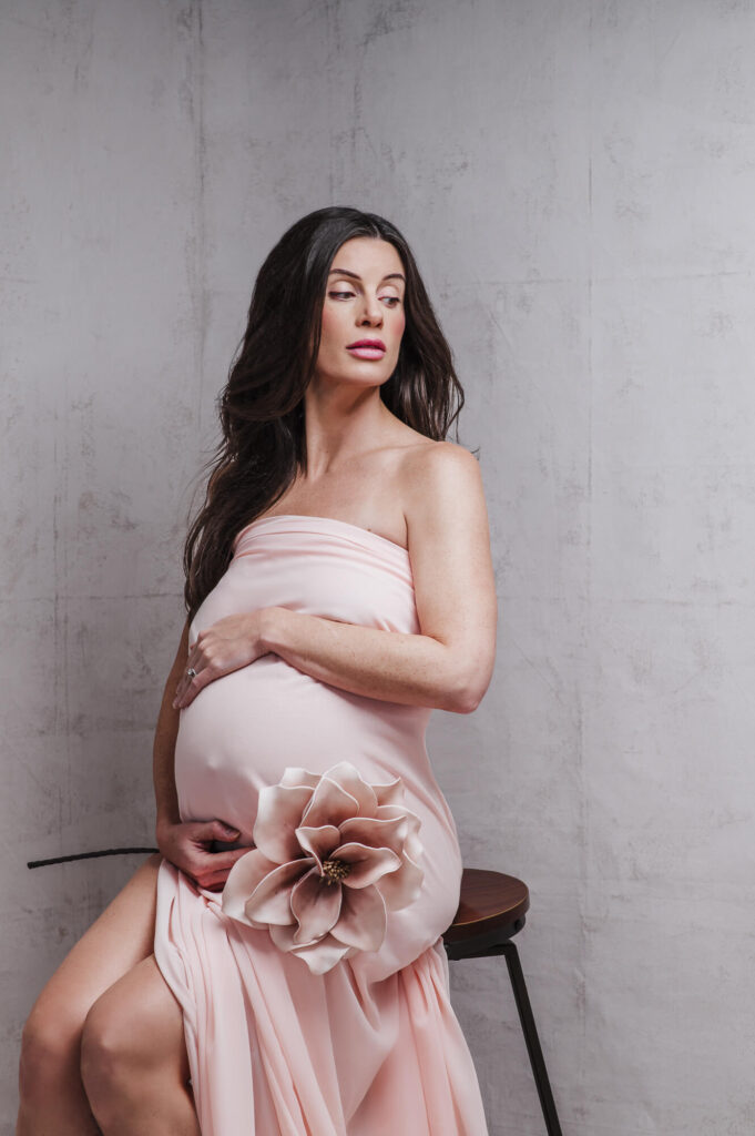 Intimate portrait of dark hair pregnant women looking sideways wrap in peach color fabric holding beautiful flower next to her belly