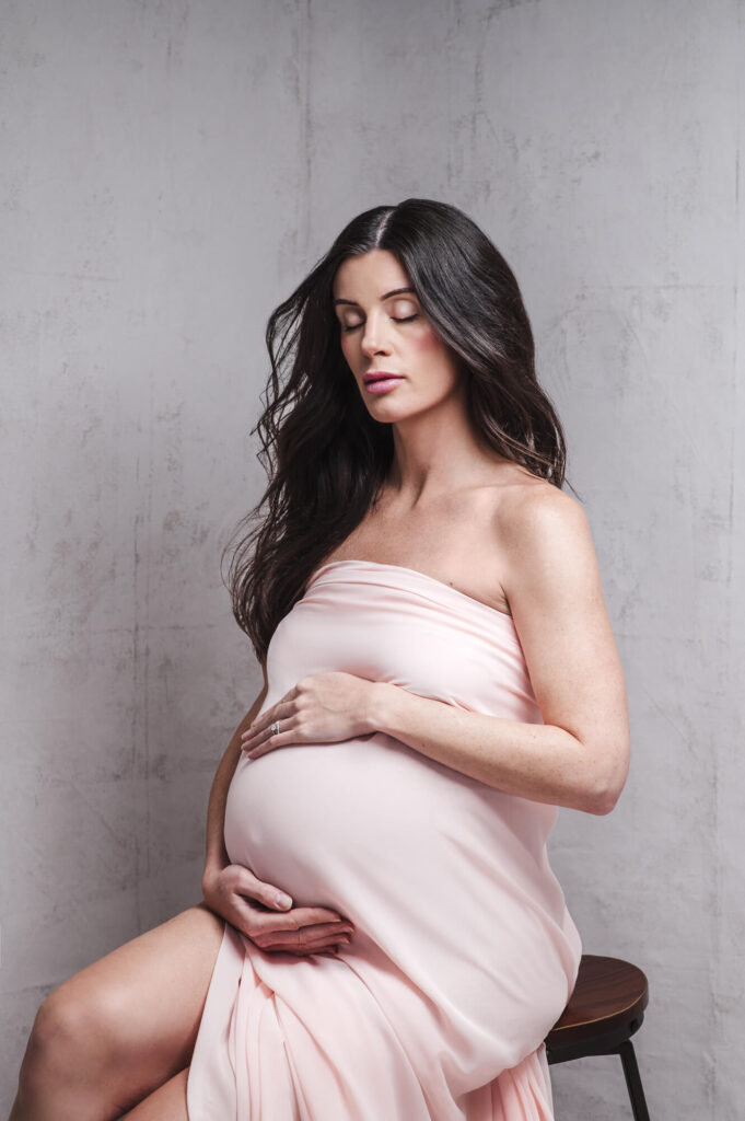 Intimate portrait of dark hair pregnant women with close eyes wrap in peach color fabric holding her belly