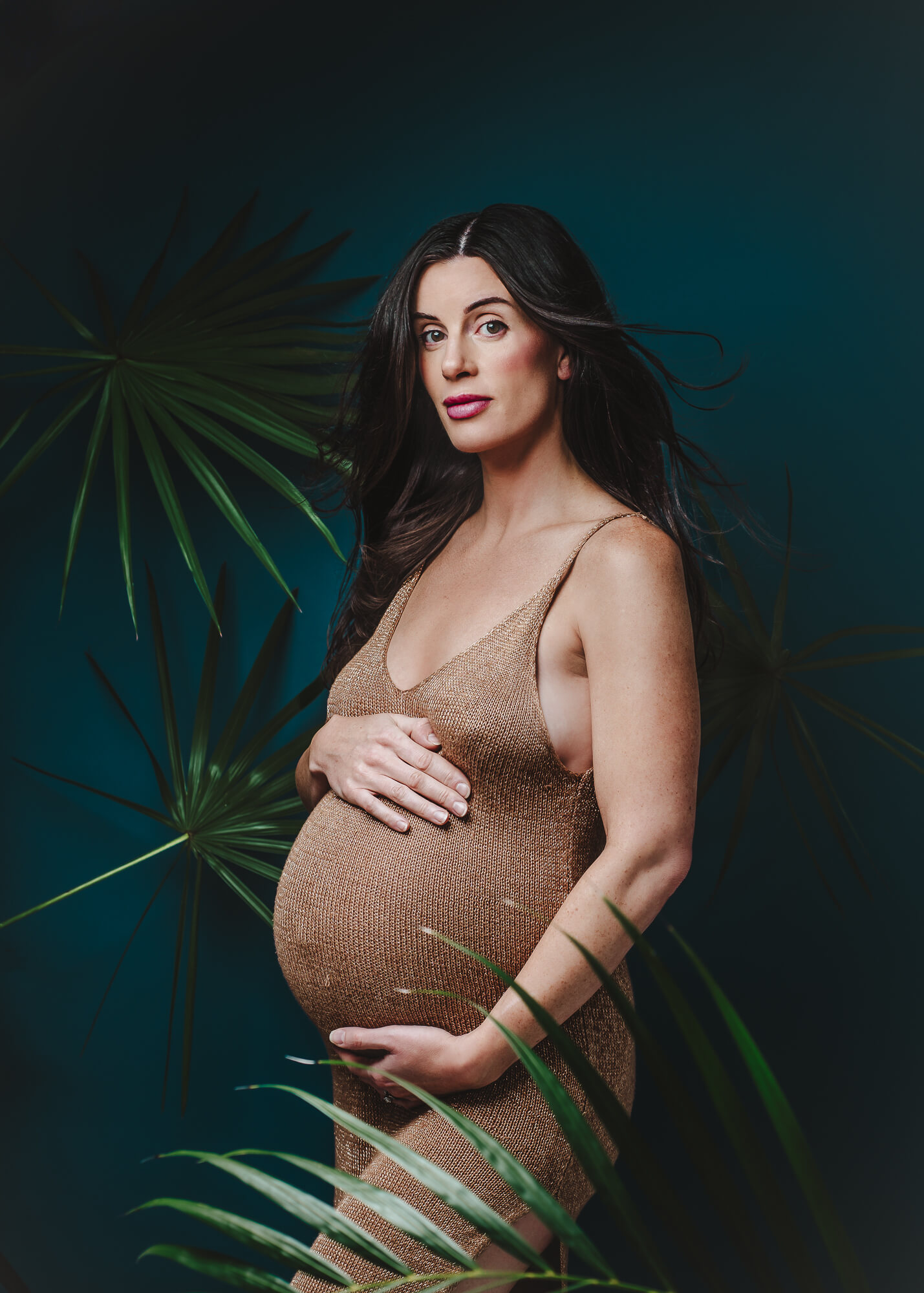 A stunning maternity client in a gold dress stands against a dark blue background, complemented by lush green palm leaves.